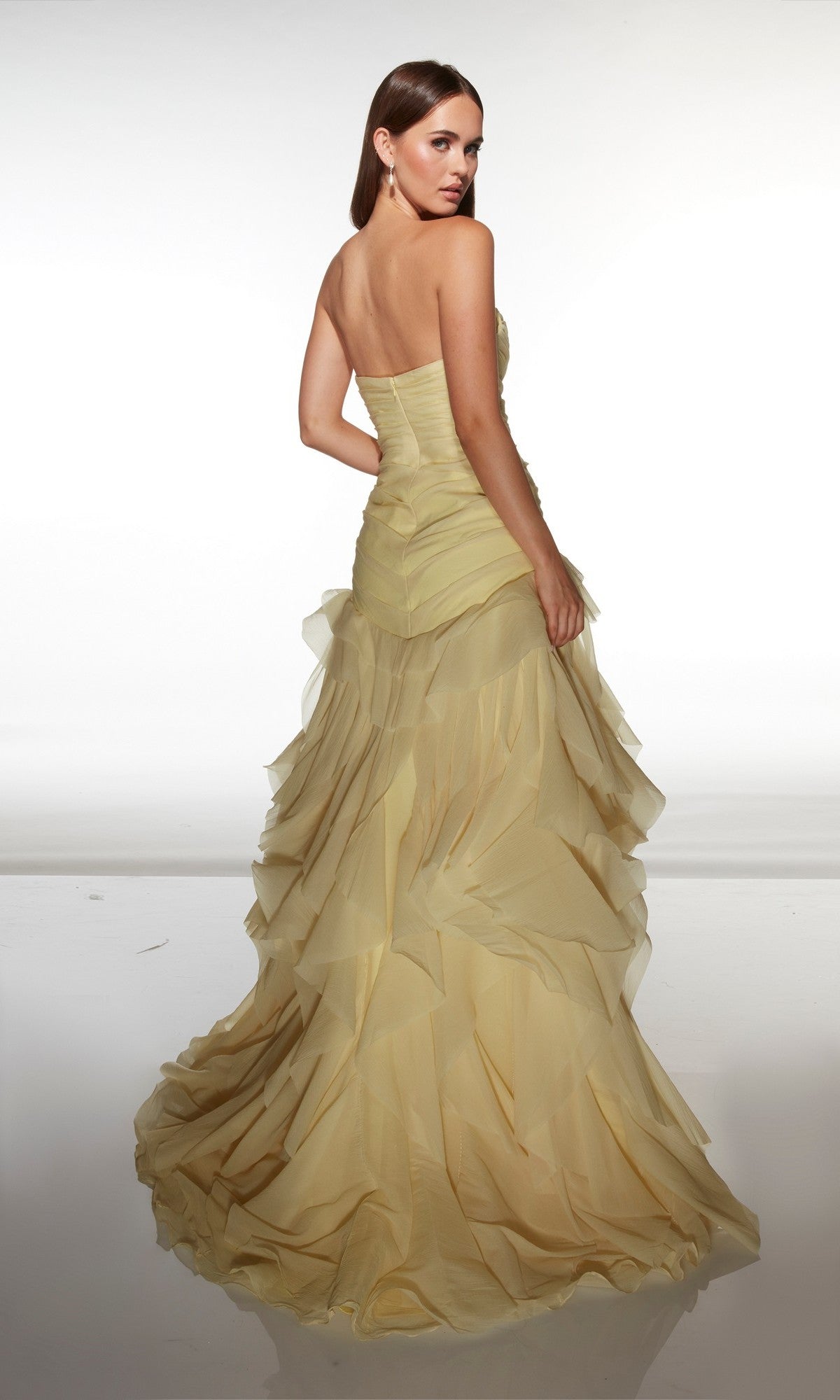 A woman faces away, wearing the Alyce 62024 long prom dress—strapless, pale yellow, with layered ruffles and a fitted bodice—set against a plain white background.